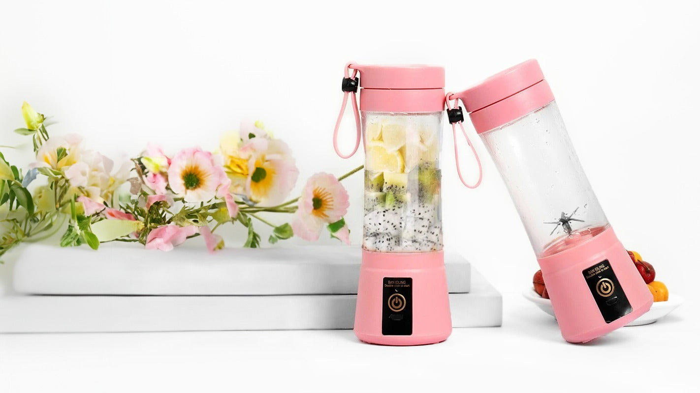 Deux blenders portables de couleur rose avec une section transparente au milieu. Le blender debout contient des fruits coupés tels que kiwi, fruit du dragon et pomme. Le second blender est incliné et vide. Ils ont tous les deux un bouton noir marqué 'GAN DING DOUBLE POWER'. Une composition de fleurs pastel décore le côté gauche de l'image, contribuant à l'esthétique de la présentation du produit.